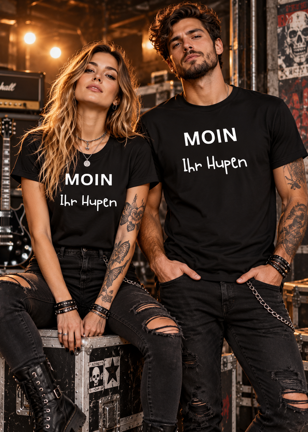 Frau und Mann im rockigen Style tragen schwarzes Unisex T-Shirt mit weißem Schriftzug MOIN Ihr Hupen vor einem industriellen Backstage-Hintergrund.