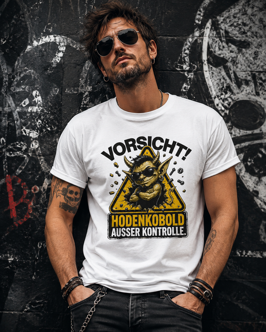 Rockig gekleideter Mann trägt ein weißes Herren T-Shirt mit gelbem Hodenkobold-Warnschild und schwarzer Jeans vor einer Graffitiwand.