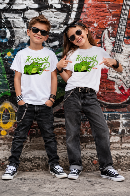 Junge und Mädchen tragen weiße Kinder T-Shirts mit grünem Krokodil-Power-Motiv vor einer bunten Graffitiwand.