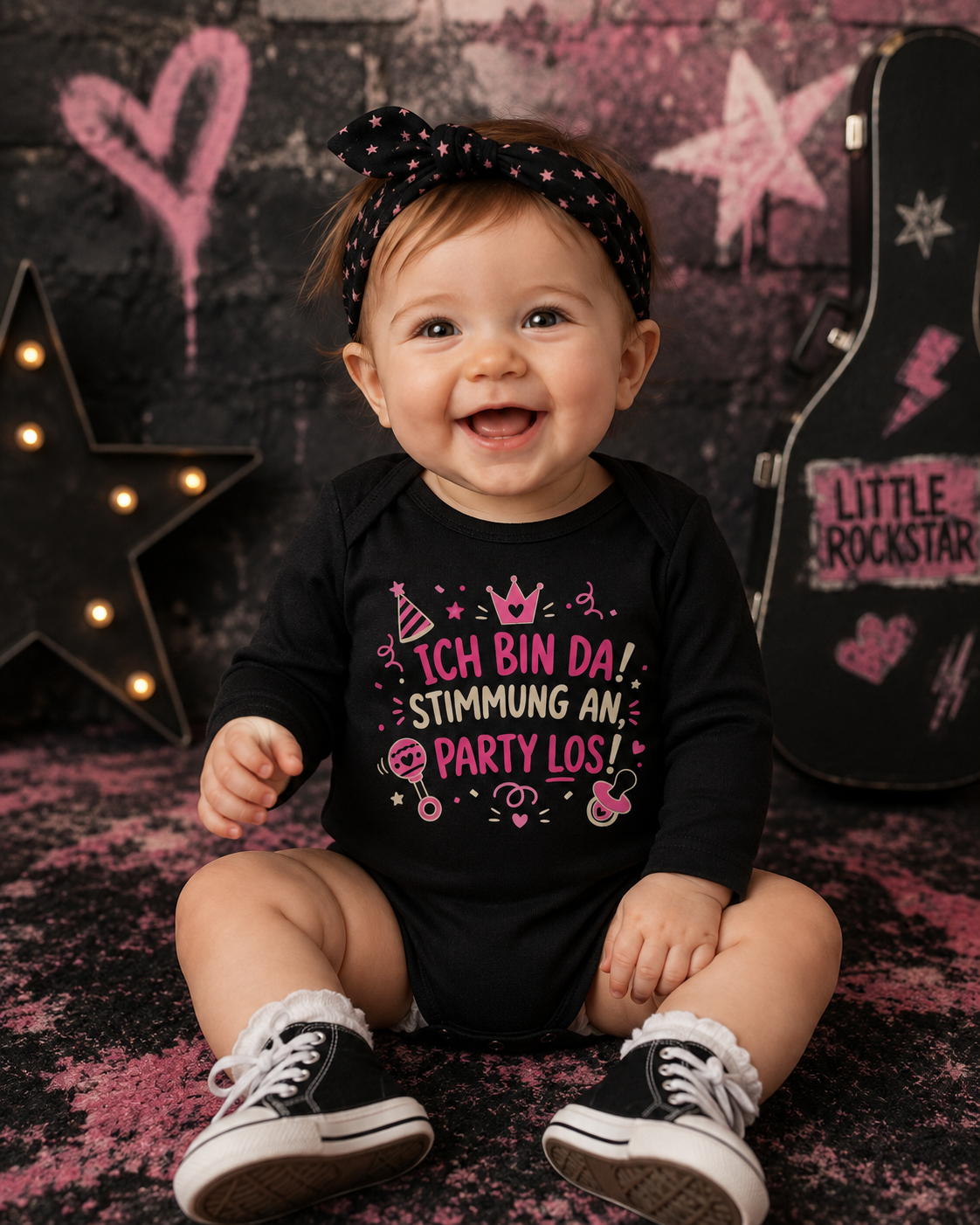 Lachendes Baby Mädchen trägt schwarzen Langarm-Babybody mit dem Spruch Ich bin da Stimmung an Party los vor rockigem Hintergrund.